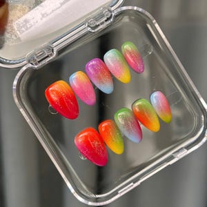 Peut inclure: Un ensemble de dix faux ongles disposés dans un boîtier de rangement en plastique transparent. Chaque ongle présente un dégradé multicolore vibrant passant par des tons de rouge, orange, jaune, vert, bleu et violet, avec des paillettes.