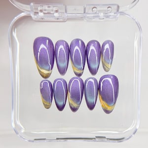 May include: A set of ten almond-shaped press-on nails in a clear plastic storage case. Each nail features a deep purple cat-eye magnetic polish finish with a swirling golden-yellow accent at the base, creating a reflective and dimensional effect.