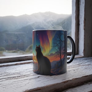 Op de afbeelding: Een zwarte keramische mok met een kleurrijke aurora borealis-scène en een zwart katten silhouet. De mok is gevuld met een stomende drank.