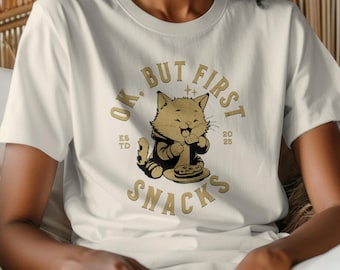 Könnte beinhalten: Ein weißes T-Shirt mit einer goldenen Grafik eines Katers, der Pizza isst. Die Grafik lautet "Ok, But First Snacks" mit dem Text "Est. 2025" unter dem Kater.