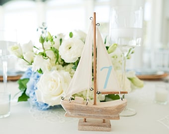 May include: A wooden sailboat table decoration with a white sail and the number 7 in light blue. The boat sits on a square base, surrounded by white and blue flowers, and is part of a table setting.
