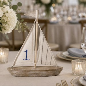 May include: A wooden sailboat table decoration with a cream-colored sail and a small flag. The sail has blue vertical stripes and the number 1. The boat is on a table with other decorations.