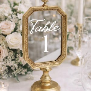 May include: Gold-colored table number holder with a mirrored surface. The text "Table 1" is displayed in white script. The holder has an octagonal frame and a pedestal base, suitable for weddings or events.