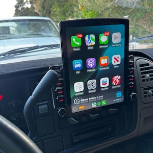 May include: A black car dashboard with a large screen displaying the iPhone home screen. The screen shows icons for Phone, Maps, Messages, Now Playing, Car, Podcasts, Audiobooks, News, Calendar, Settings, Chevron, Chick-fil-A, and a notification that the iPhone battery is low. The screen is mounted in a black frame.