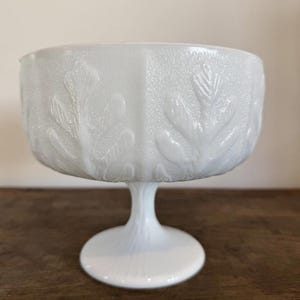 Könnte beinhalten: Eine weiße Milchglas-Fußschale mit floralem Muster. Die Schale hat einen breiten, runden Boden und einen hohen, schlanken Stiel.