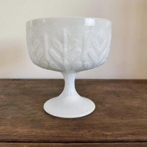 Puede incluir: Tazón de pedestal de vidrio de leche blanco con un diseño de hojas en relieve. El tazón está sobre una superficie de madera.