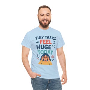 May include: Light blue t-shirt with the text "TINY TASKS = FEEL HUGE TODAY" in navy blue, red, and teal. The shirt features a graphic of a person with their hands on their head. The person is wearing a yellow shirt.
