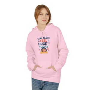 Può includere: Una felpa con cappuccio rosa chiaro con una tasca frontale e un cappuccio con coulisse. La felpa con cappuccio presenta il testo "TINY TASKS FEEL HUGE TODAY" con una grafica di una persona con le mani sulla testa. La felpa è realizzata in materiale morbido.