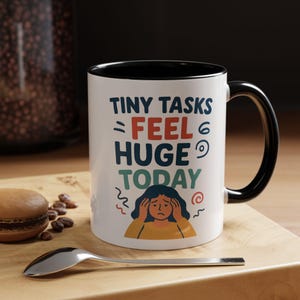 May include: White ceramic mug with a black interior and handle. The mug features the text "TINY TASKS FEEL HUGE TODAY" in various colors, along with an illustration of a person holding their head. A spoon, macaron, and coffee beans are on a wooden surface.
