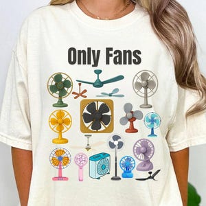 May include: Cream-colored t-shirt with the text "Only Fans" in black. The shirt features a colorful graphic of various fans, including ceiling fans, desk fans, and handheld fans. The fans are in various colors and styles.