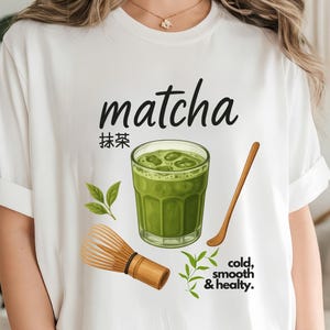 Puede incluir: Camiseta blanca con un diseño de matcha. El diseño incluye la palabra "matcha" en escritura negra, caracteres japoneses, un vaso de matcha verde con hielo, una cuchara de madera, un batidor de bambú y el texto "cold, smooth & healty."
