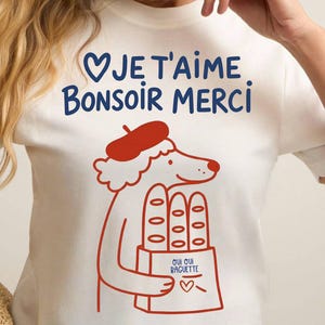 Puede incluir: Camiseta blanca con una ilustración roja y azul de un perro con boina y sosteniendo una bolsa de baguettes. El texto "Je t'aime. Bonsoir Merci" está impreso en azul en la camiseta.
