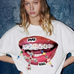 May include: White t-shirt featuring a graphic of a mouth with braces and the words "SAVAGE" and "REBEL". The mouth graphic has colorful gems and a cross pendant. The model is wearing a blue skirt.