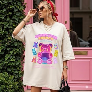 Pode incluir: T-shirt bege oversized com um gráfico colorido de um urso de goma e o texto "Gummy Bear Energy". O design inclui várias ilustrações de ursos de goma em diferentes cores. A camisa tem mangas curtas.