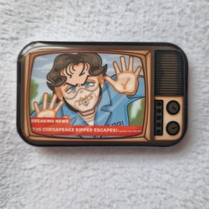 Puede incluir: Pin rectangular con una imagen de dibujos animados de un hombre en una pantalla de televisión. El hombre lleva una camisa azul y tiene las manos en alto. El texto en la pantalla dice "BREAKING NEWS THE CHESAPEAKE RIPPER ESCAPES!"