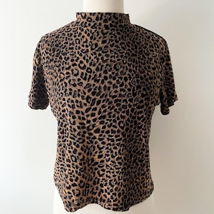 May include: A short-sleeved top with a mock turtleneck, featuring a brown and black leopard print pattern. The shirt is displayed on a white mannequin, showcasing its design and fit. The fabric appears soft and lightweight.