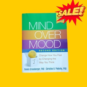 May include: A book titled "Mind Over Mood: Second Edition" by Dennis Greenberger, PhD and Christine A. Padesky, PhD. The book cover features a yellow smiley face in a box with a frowning face on the bottom of the box. The book cover is a multi-colored design with the title in white text.