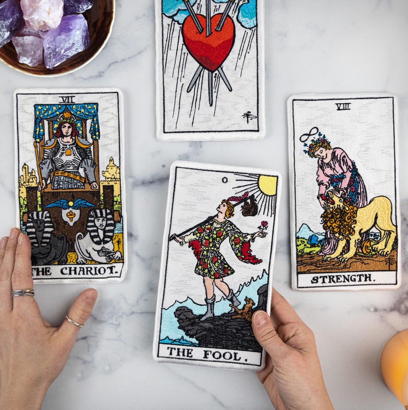 May include: Four tarot card patches with embroidered designs. The cards include "The Chariot," "Three of Swords," "Strength," and "The Fool." The cards are white with colorful illustrations and black text. The cards are on a white marble surface.