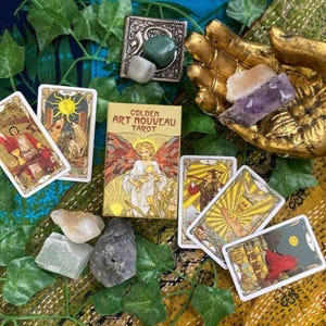 May include: A Golden Art Nouveau Tarot deck with several tarot cards fanned out. The deck box and cards feature illustrations of figures and symbols. The image also includes crystals, a gold hand statue, and decorative greenery.