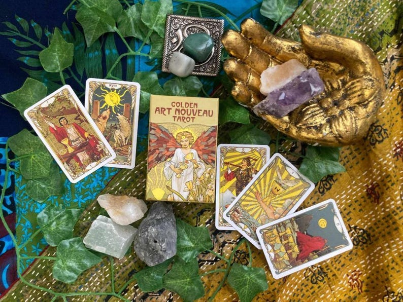 May include: A Golden Art Nouveau Tarot deck with several tarot cards fanned out. The deck box and cards feature illustrations of figures and symbols. The image also includes crystals, a gold hand statue, and decorative greenery.