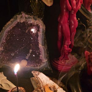 May include: A still life arrangement featuring a large amethyst geode, a red candle shaped like embracing figures, and a burning incense stick. Other elements include a seashell, a nautilus shell, and dried flowers.