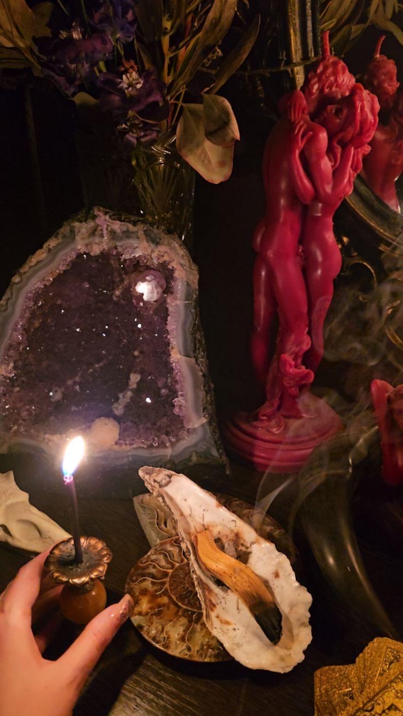 May include: A still life arrangement featuring a large amethyst geode, a red candle shaped like embracing figures, and a burning incense stick. Other elements include a seashell, a nautilus shell, and dried flowers.