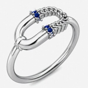 May include: A silver ring with a distinctive design. The ring has a chain-style band and two curved sections, each set with small blue gemstones. The ring's design is modern and elegant, suitable for everyday wear or special occasions.