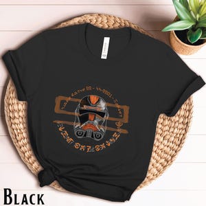 Może przedstawiać: Czarny T-shirt z grafiką pomarańczowego i białego hełmu i tekstem w okrągłym wzorze. Tekst brzmi "The Bad Batch" w stylizowanym foncie.