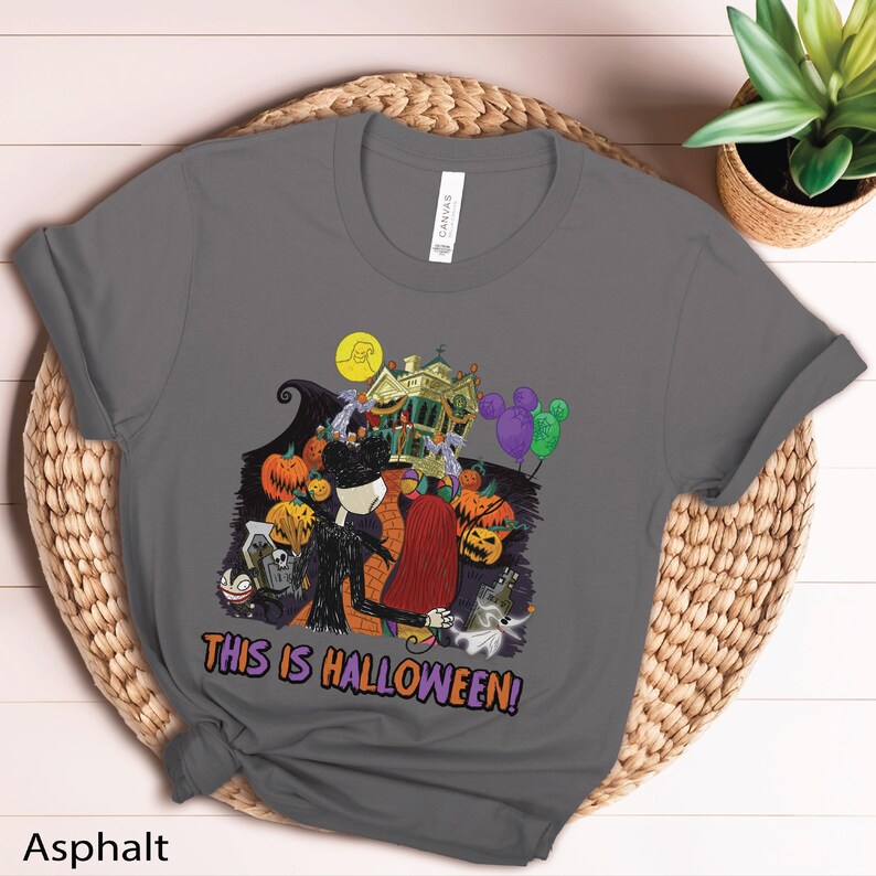 Puede incluir: Camiseta gris asfalto con un dise&ntilde;o de Halloween. El dise&ntilde;o presenta una casa embrujada, calabazas y personajes, con el texto "THIS IS HALLOWEEN!". La camiseta est&aacute; hecha de un material suave.