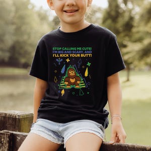 May include: A black t-shirt with a green, yellow, and blue graphic of a Bigfoot character. The text on the shirt reads "STOP CALLING ME CUTE! I'M BIG AND SCARY, AND I'LL KICK YOUR BUTT!"