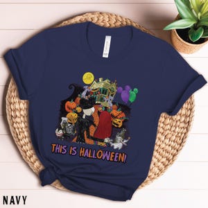 Puede incluir: Camiseta azul marino con un gr&aacute;fico colorido de Halloween. El dise&ntilde;o incluye a Jack Skellington y Sally, calabazas, una casa embrujada y el texto "THIS IS HALLOWEEN!". La camiseta se muestra sobre una superficie tejida.