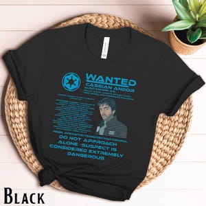 May include: A black t-shirt with a blue and white wanted poster design featuring a character from the Star Wars universe. The poster reads "WANTED CASSIAN ANDOR ALSO KNOWN AS: WILLIK AACH, JOBETH SWARD, CLEM, KEEF SIC, AND CODENAME PULCRUM". The poster also includes a warning: "DO NOT APPROACH ALONE - SUSPECT IS CONSIDERED EXTREMELY DANGEROUS".