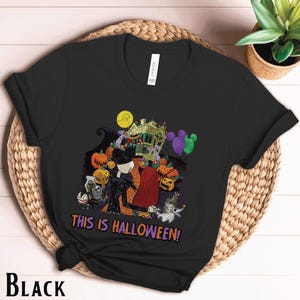 Puede incluir: Camiseta negra con un gr&aacute;fico colorido de Halloween. El dise&ntilde;o incluye personajes, calabazas y una casa embrujada, con el texto "THIS IS HALLOWEEN!". La camiseta se muestra sobre una superficie tejida.