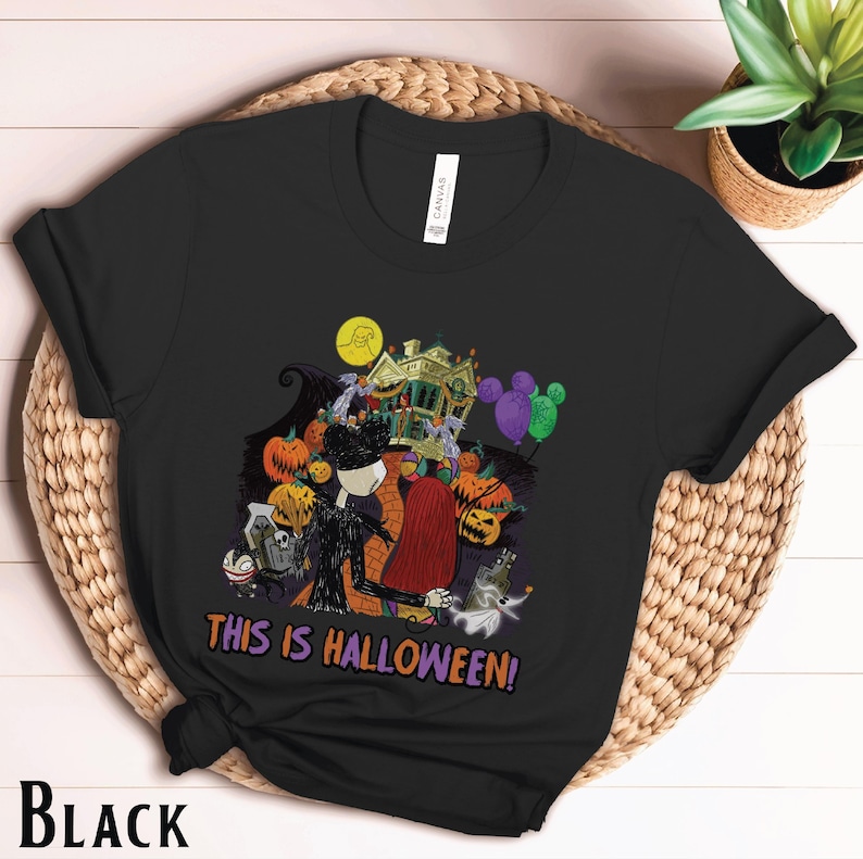 Puede incluir: Camiseta negra con un gr&aacute;fico colorido de Halloween. El dise&ntilde;o incluye personajes, calabazas y una casa embrujada, con el texto "THIS IS HALLOWEEN!". La camiseta se muestra sobre una superficie tejida.