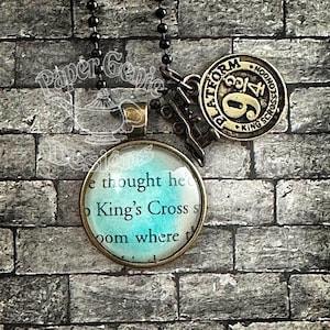 May include: A black chain necklace with three pendants. One pendant is a round, bronze-colored charm with the text "Platform 9 3/4 King's Cross London". Another pendant is a bronze-colored train. The third pendant is a round, glass-covered charm with text.