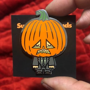 Puede incluir: Un pin de metal con una calabaza de dibujos animados con una cara triste que lleva un traje y una corbata. El pin está sobre un fondo negro con el texto "Super Friends" en blanco.