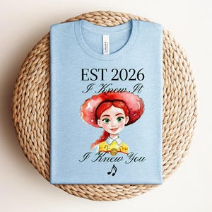 Pode incluir: Uma t-shirt azul-clara com uma ilustra&ccedil;&atilde;o em estilo aguarela de uma personagem ruiva com um chap&eacute;u vermelho grande. O texto diz EST 2026, I Knew It e I Knew You, acompanhado por uma pequena nota musical preta abaixo do texto.