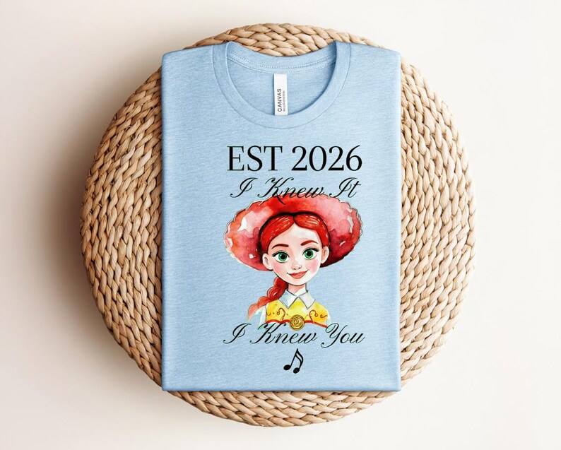 Pode incluir: Uma t-shirt azul-clara com uma ilustra&ccedil;&atilde;o em estilo aguarela de uma personagem ruiva com um chap&eacute;u vermelho grande. O texto diz EST 2026, I Knew It e I Knew You, acompanhado por uma pequena nota musical preta abaixo do texto.