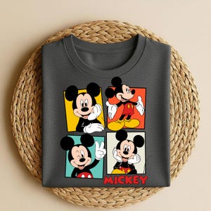以下が含まれることがあります： ミッキーマウスのイラストが描かれた4つのパネルが付いた濃いグレーのTシャツ。各パネルには異なるポーズと背景色（黄色、赤、青、ベージュ）があります。「MICKEY」の文字が赤色で下部に印刷されています。