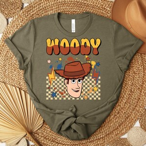 Puede incluir: Camiseta verde oliva con la palabra "WOODY" en fuente retro naranja y marr&oacute;n. La camiseta presenta una ilustraci&oacute;n de dibujos animados de Woody con un sombrero de vaquero marr&oacute;n, estrellas y rayos. Un sombrero de vaquero marr&oacute;n es visible en el fondo.