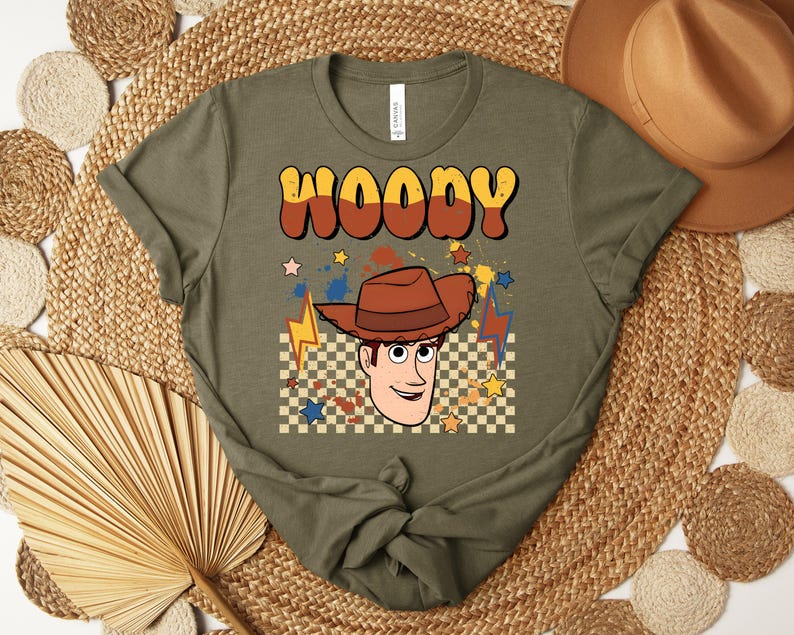 Puede incluir: Camiseta verde oliva con la palabra "WOODY" en fuente retro naranja y marr&oacute;n. La camiseta presenta una ilustraci&oacute;n de dibujos animados de Woody con un sombrero de vaquero marr&oacute;n, estrellas y rayos. Un sombrero de vaquero marr&oacute;n es visible en el fondo.