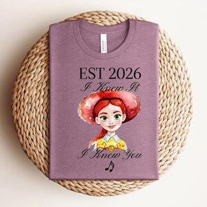 Pode incluir: Uma t-shirt malva com uma ilustra&ccedil;&atilde;o em estilo aguarela de uma personagem ruiva com um chap&eacute;u vermelho. O texto diz EST 2026, I Knew It e I Knew You, acompanhado por um pequeno s&iacute;mbolo de nota musical impresso em tinta preta.