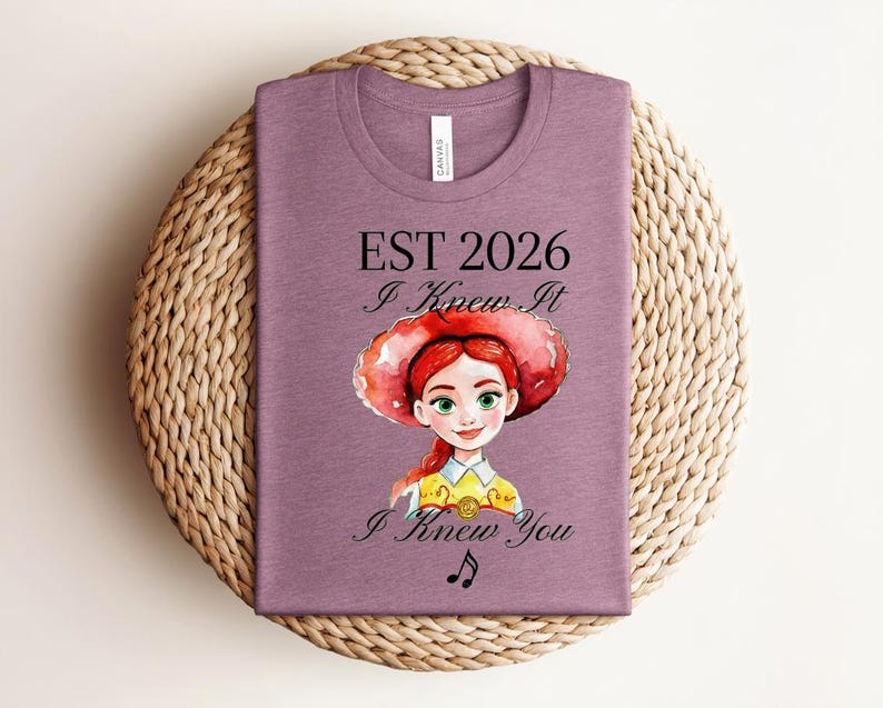 Pode incluir: Uma t-shirt malva com uma ilustra&ccedil;&atilde;o em estilo aguarela de uma personagem ruiva com um chap&eacute;u vermelho. O texto diz EST 2026, I Knew It e I Knew You, acompanhado por um pequeno s&iacute;mbolo de nota musical impresso em tinta preta.