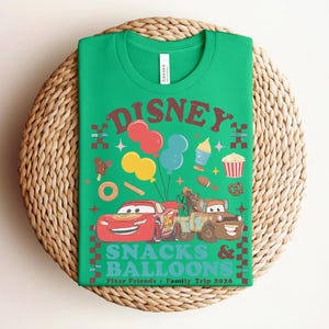 May include: Green t-shirt with a graphic featuring the word "DISNEY" in red, balloons, and cartoon cars. The text "SNACKS & BALLOONS" is below the cars. The shirt is on a woven, circular surface.