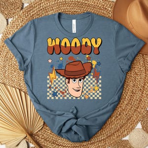 Puede incluir: Una camiseta azul con la palabra "WOODY" en fuente retro marr&oacute;n y amarilla. Debajo del texto hay una imagen de dibujos animados de Woody con un sombrero de vaquero marr&oacute;n, rodeado de estrellas y rayos. La camiseta es de material suave.