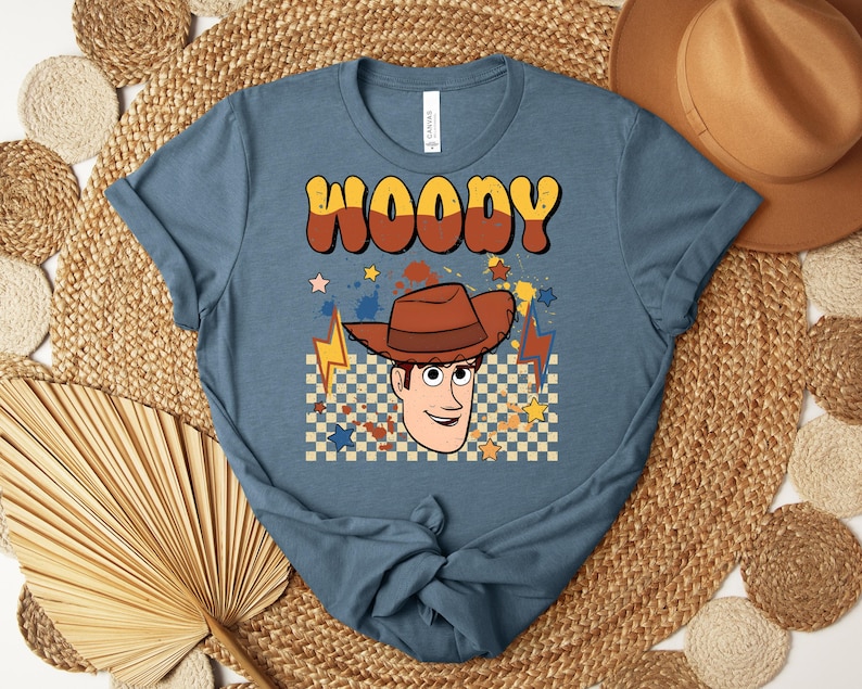 Puede incluir: Una camiseta azul con la palabra "WOODY" en fuente retro marr&oacute;n y amarilla. Debajo del texto hay una imagen de dibujos animados de Woody con un sombrero de vaquero marr&oacute;n, rodeado de estrellas y rayos. La camiseta es de material suave.