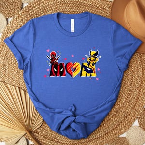 Op de afbeelding: Blauw t-shirt met het woord "MOM" en Deadpool- en Wolverine-afbeeldingen. De "M" is in de kleuren van Deadpool, de "O" is een hart en de "M" is in de kleuren van Wolverine. Kleine roze hartjes zijn verspreid over het ontwerp.