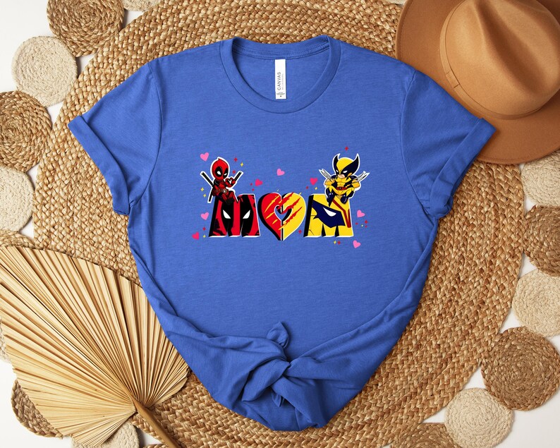 Op de afbeelding: Blauw t-shirt met het woord "MOM" en Deadpool- en Wolverine-afbeeldingen. De "M" is in de kleuren van Deadpool, de "O" is een hart en de "M" is in de kleuren van Wolverine. Kleine roze hartjes zijn verspreid over het ontwerp.