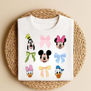 May include: White t-shirt with cartoon character faces and bows. The shirt features images of Mickey Mouse, Minnie Mouse, Donald Duck, Daisy Duck, and Goofy, alongside various colourful bows. The shirt is folded on a woven, circular mat.
