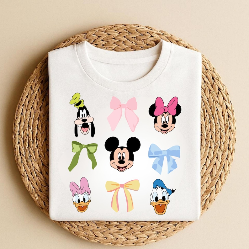 May include: White t-shirt with cartoon character faces and bows. The shirt features images of Mickey Mouse, Minnie Mouse, Donald Duck, Daisy Duck, and Goofy, alongside various colourful bows. The shirt is folded on a woven, circular mat.